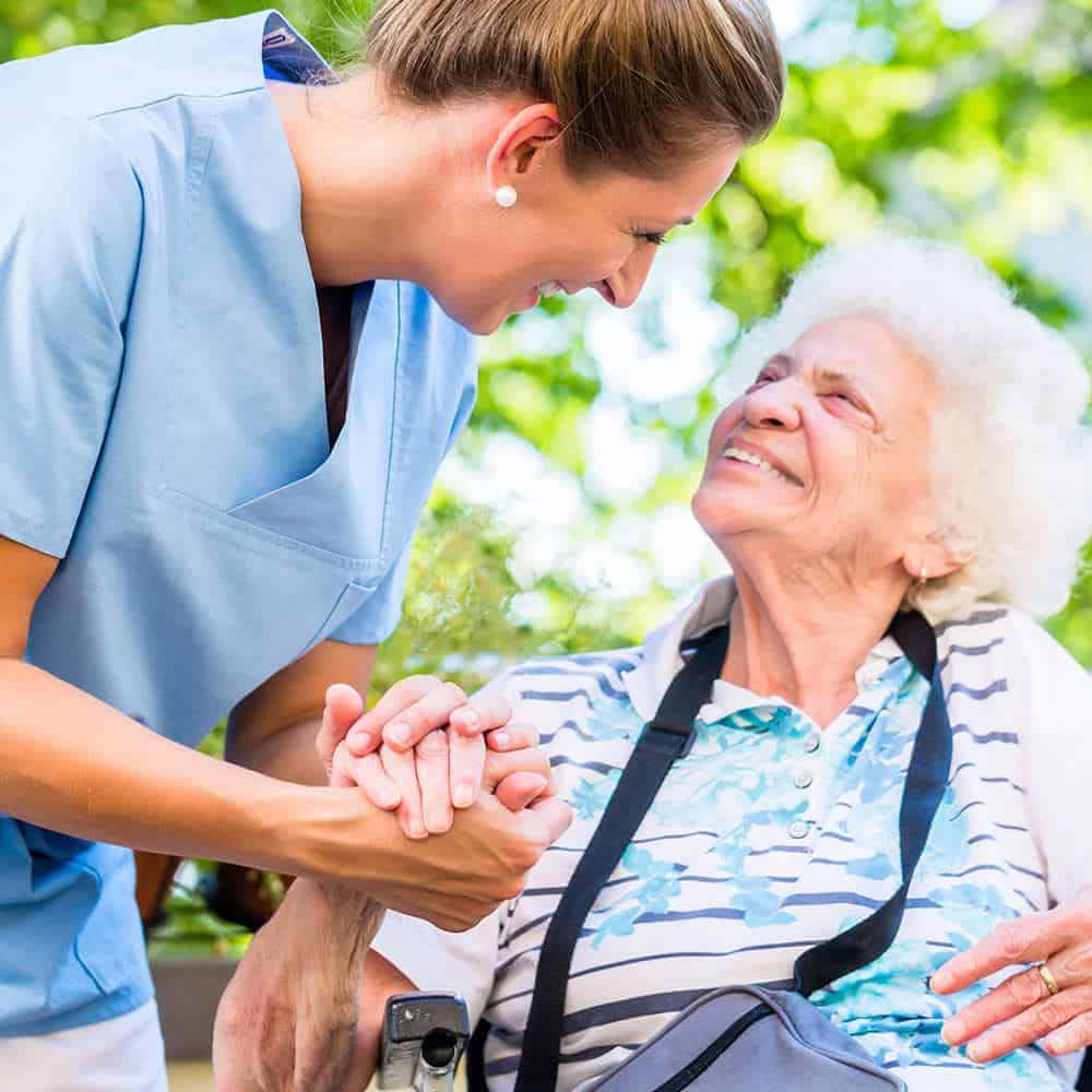 Intensivpflege Recklinghausen - Seniorin im Rollstuhl und Pflegerin halten sich die Hände und lächeln glücklich